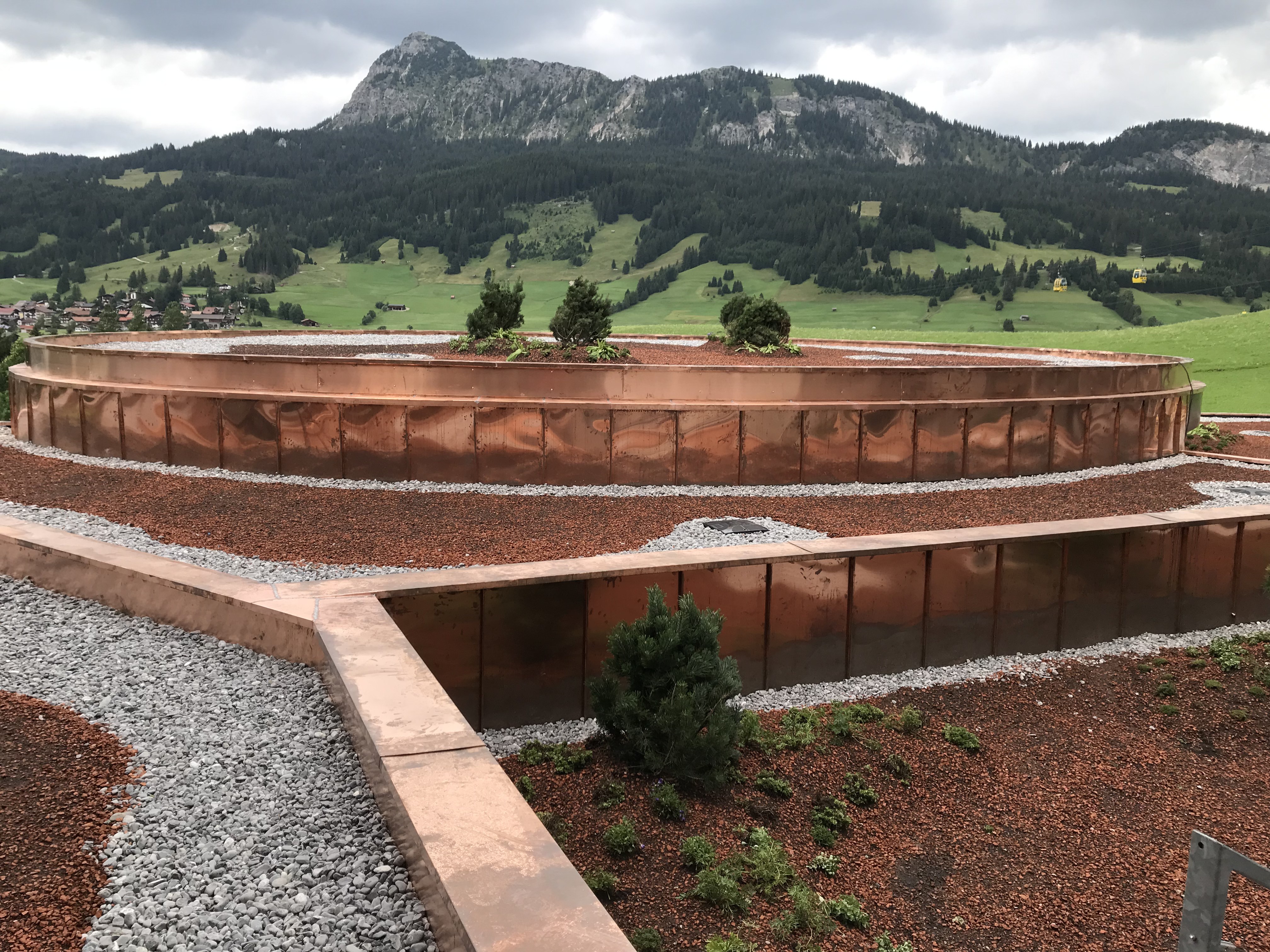 Kupferner Dachgarten mit kleinen Sträuchern, umgeben von Bergen unter einem bewölkten Himmel. Die Szene wirkt ruhig und natürlich, mit erdigen Farbtönen.